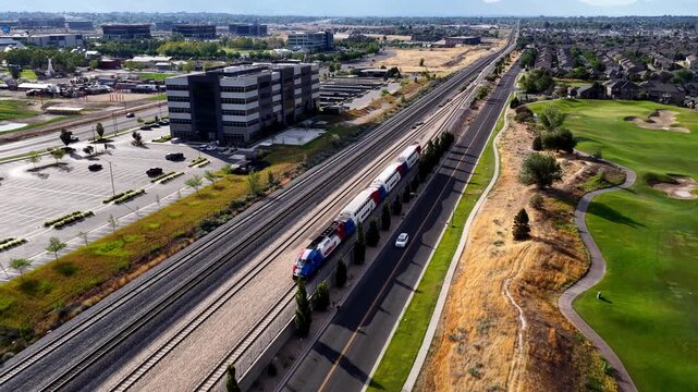 Aerial view of a commuter train moving along tracks next to an office building with solar panels and a solar panel covered parking area in Lehi, Utah, United States.