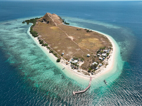 Aerial view of Pulau Kenawa with its grassy hills, sandy beaches, turquoise coral reefs, and a wooden pier extending into the clear water in Poto Tano, West Nusa Tenggara, Indonesia.