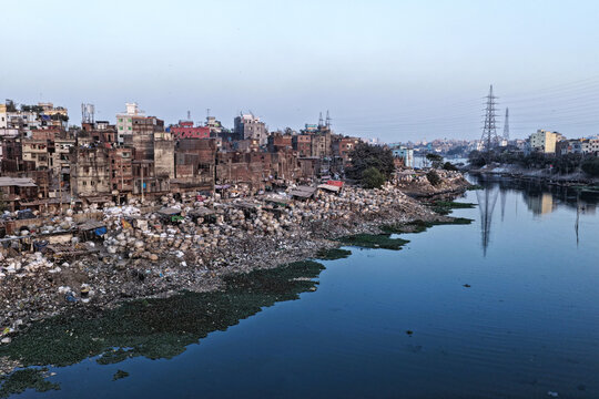 Aerial view of the Buriganga River flowing past dense urban buildings and a cluttered riverbank with debris and water hyacinth in Dhaka, Dhaka Division, Bangladesh.