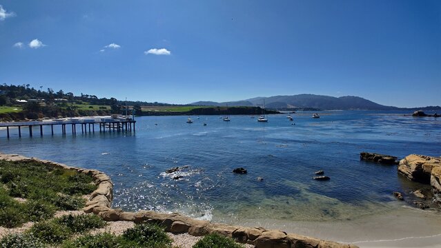 View of 17 Mile Drive featuring a wooden pier extending into the calm bay with sailboats, rocky shoreline, and green hills in Del Monte Forest, California, United States.