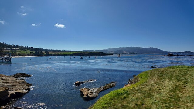 View of 17 Mile Drive with boats anchored in the calm bay, rocky coastline, and green grass under a clear blue sky in Del Monte Forest, California, United States.