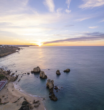 Aerial view of Praia dos Arrifes with its distinctive rock formations rising from the calm sea, sandy beach, and golden sunset light in Faro District, Portugal.