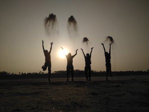 View of silhouettes of four people jumping and throwing sand into the air against a bright sunset sky on a riverbank in Kushtia, Khulna Division, Bangladesh.