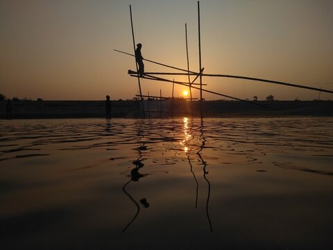 View of a fisherman standing on a bamboo fishing structure in the river at sunset in Kushtia, Khulna Division, Bangladesh.