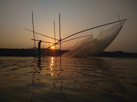 View of a fisherman operating a traditional bamboo lift net on the river at sunset with golden light reflecting on the water in Kushtia, Khulna Division, Bangladesh.