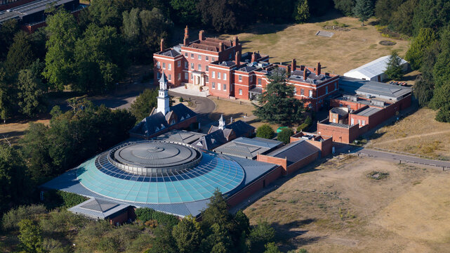 Aerial view of the Wellcome Genome Campus featuring a historic red brick building and a modern circular glass-domed structure surrounded by lush parkland in Hinxton, England, United Kingdom.