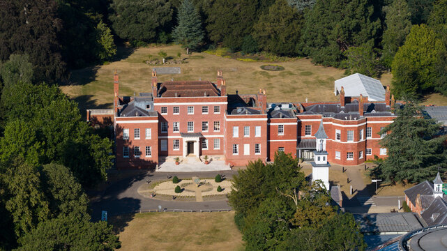 Aerial view of Hinxton Hall at the Wellcome Genome Campus, featuring the historic red brick mansion surrounded by lush green trees and manicured grounds in Hinxton, England, United Kingdom.