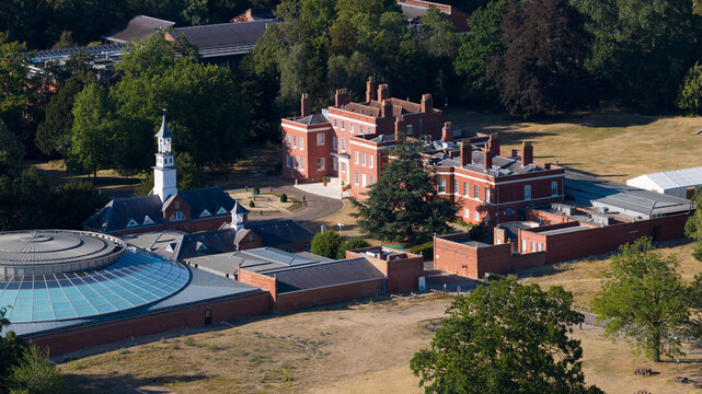 Aerial view of the Wellcome Genome Campus featuring historic red brick buildings, a modern circular glass-roofed structure, and surrounding parkland in Hinxton, England, United Kingdom.