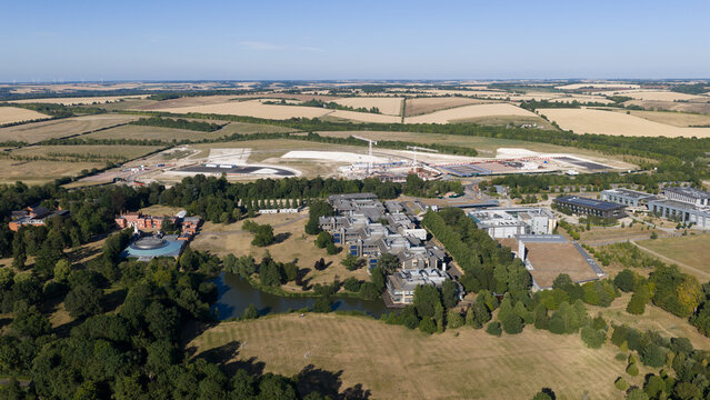 Aerial view of Wellcome Genome Campus featuring modern research buildings, a small lake, surrounding green spaces, and construction areas in Hinxton, England, United Kingdom.