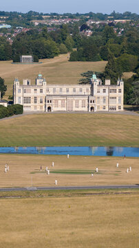 Aerial view of Audley End House surrounded by expansive parkland with a cricket match in progress on the lawn in Audley End, England, United Kingdom.