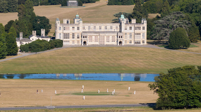 Aerial view of Audley End House with a cricket match being played on the lawn in the foreground, surrounded by expansive parkland and trees in Audley End, England, United Kingdom.