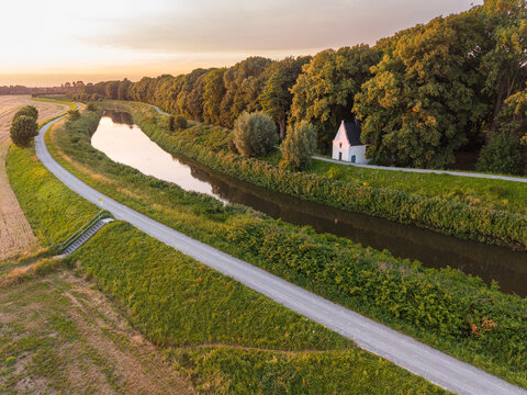 Aerial view of Kapel O-L-Vrouw in 't Hammeke situated along the Zenne river surrounded by lush trees and rural fields during a golden sunset in Zemst, Flanders, Belgium.