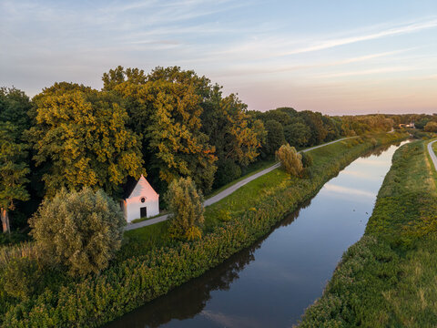 Aerial view of Kapel O-L-Vrouw in 't Hammeke nestled among lush trees beside the Zenne river during a golden sunset in Zemst, Flanders, Belgium.