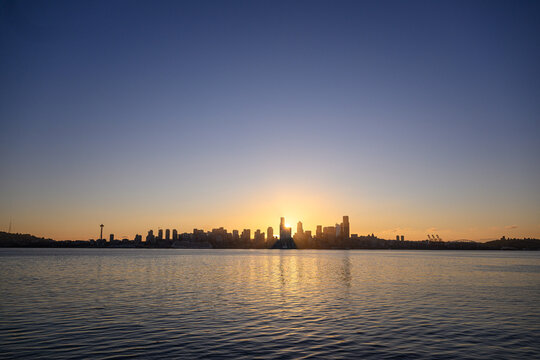 View of the Seattle skyline at sunset with the sun behind skyscrapers, reflecting on the calm waters of Elliott Bay, seen from Don Armeni Boat Ramp in Seattle, Washington, United States.