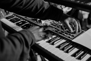 Black and white close-up of a producer hands tweaking dials on a music synthesizer keyboard. © Caleb