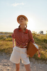 Obraz premium Smiling woman in striped shirt and white shorts wearing a straw hat, standing outdoor under warm sunshine. Casual summer lifestyle and travel vibe, confident and bright.