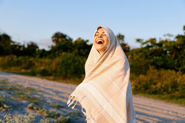 Obraz premium Young woman wrapped in a light blanket laughs outdoors on a sunny beach path, radiating joy, wellness, and carefree summer vibes.