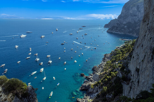 Aerial view of Capri with numerous boats and yachts anchored in the turquoise bay, surrounded by dramatic steep cliffs and lush greenery in Capri, Campania, Italy.