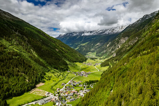 Aerial view of the village of Prutz nestled in a lush green valley surrounded by steep, pine-covered mountains and snow-capped peaks under a cloudy sky in Prutz, Tyrol, Austria.