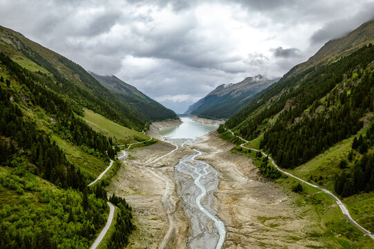 Aerial view of the mountain valley with a winding river flowing into a reservoir, surrounded by lush green pine forests and steep slopes under a cloudy sky in Prutz, Tyrol, Austria.