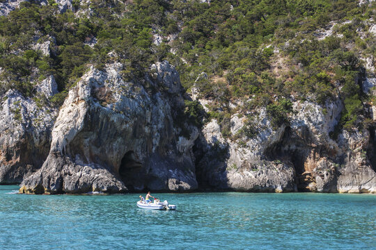 View of a small motorboat with people cruising in the turquoise waters of the Gulf of Orosei, framed by rugged limestone cliffs and Mediterranean vegetation, Italy.