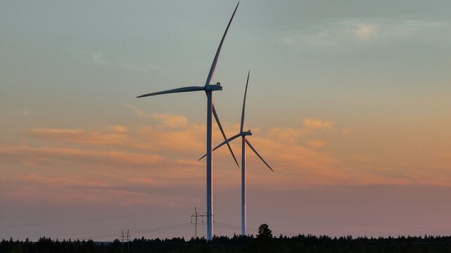Aerial view of Metsalamminkangas Tuulipuisto wind turbines standing tall against a soft golden hour sky above a dense forest landscape, Siikalatva, Pohjois-Pohjanmaa, Finland.