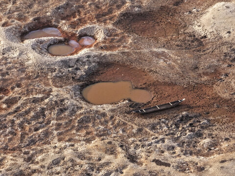 Aerial view of the dry, cracked riverbed of the Jadukata River with small stagnant pools of water and a single wooden boat resting on the parched earth, Sunamganj, Sylhet Division, Bangladesh.