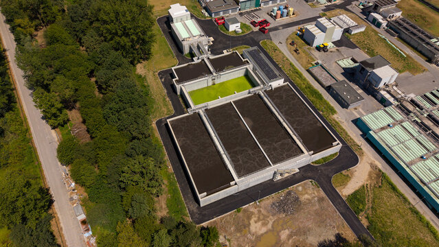 Aerial view of a wastewater treatment plant featuring large sedimentation tanks with green water, industrial infrastructure, and surrounding lush green trees in Milan, Lombardy, Italy.