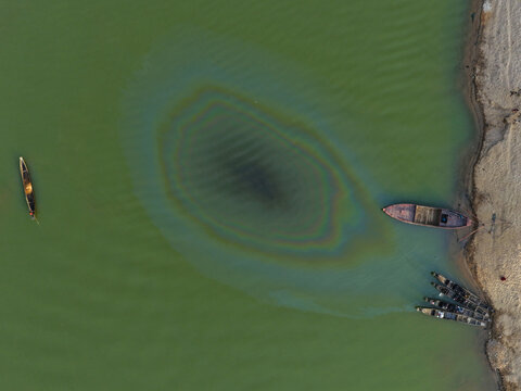 Aerial view of Jadukata River with traditional wooden boats moored along the sandy bank and an iridescent oil slick on the water surface in Sunamganj, Sylhet Division, Bangladesh.