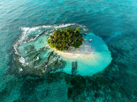Aerial view of Guyam Island featuring a small tropical island with lush palm trees, white sand beach, and clear turquoise water surrounded by coral reefs in General Luna, Caraga, Philippines.