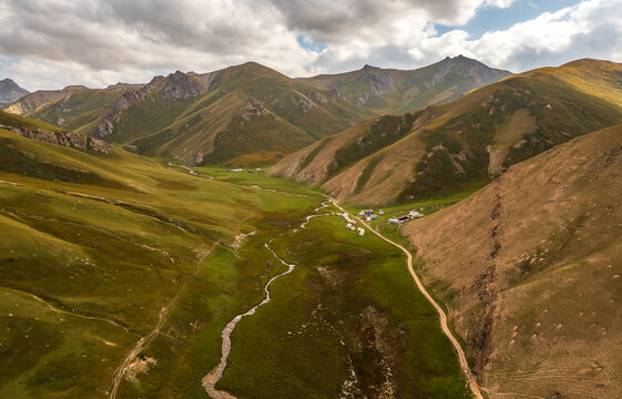 Aerial view of Tash Rabat Caravan Palace in a green valley surrounded by rugged mountains, with a winding stream and traditional yurts in At-Bashi District, Naryn Region, Kyrgyzstan.