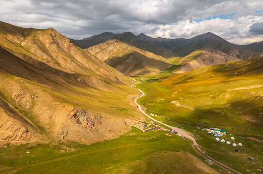 Aerial view of Tash Rabat Caravan Palace situated in a vast mountain valley with rolling green hills, a winding dirt road, and traditional yurts in At-Bashi District, Naryn Region, Kyrgyzstan.