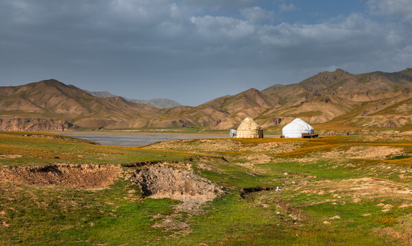View of yurts situated in a vast, grassy valley near the shores of Kol-Suu lake, surrounded by rugged mountains under a cloudy sky in At-Bashi District, Naryn Region, Kyrgyzstan.