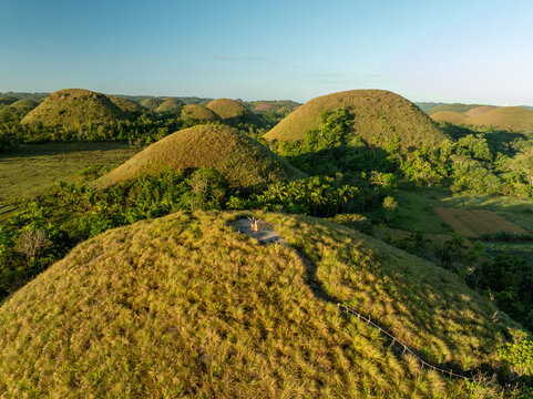 Aerial view of the Chocolate Hills with a person standing on a grassy peak, surrounded by iconic conical hills and lush tropical vegetation in Batuan, Central Visayas, Philippines.