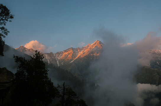 View of the Triund Trek Base Camp with golden light hitting the mountain peaks and clouds swirling around the slopes in Dharamshala, Himachal Pradesh, India.