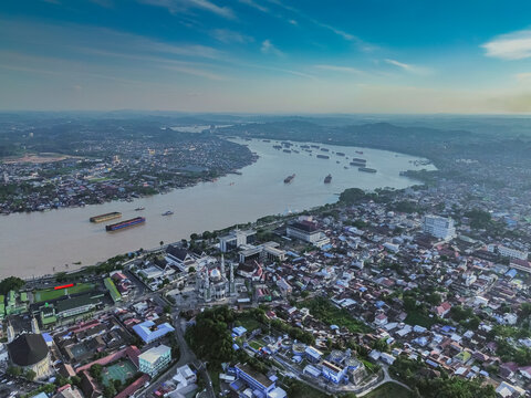 Aerial view of the Mahakam River flowing through Samarinda, featuring the Islamic Center Mosque, dense urban rooftops, and cargo barges on the water in Samarinda, Kalimantan Timur, Indonesia.
