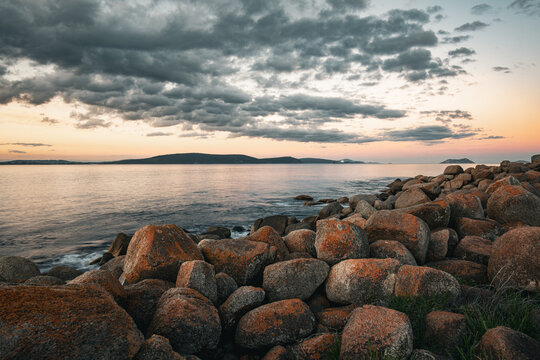 View of the rocky coastline at Middleton Beach with orange-lichen-covered boulders, calm ocean waters, and a dramatic cloudy sky at sunset in Middleton Beach, Western Australia, Australia.