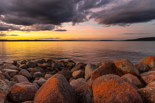 View of sunset over the ocean with large lichen-covered rocks in the foreground and dramatic dark clouds at Middleton Beach, Western Australia, Australia.