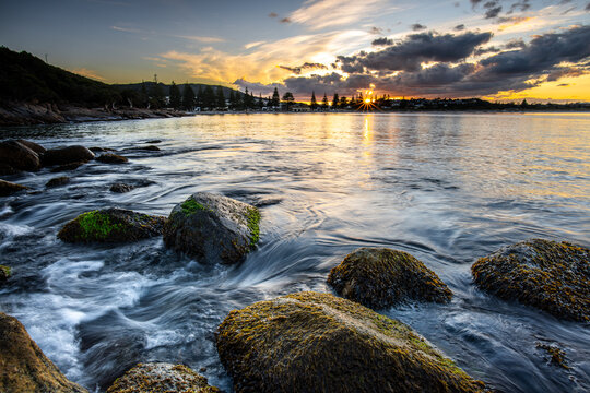 View of Middleton Beach at sunset with moss-covered rocks in the foreground and calm ocean waves under a dramatic sky in Middleton Beach, Western Australia, Australia.