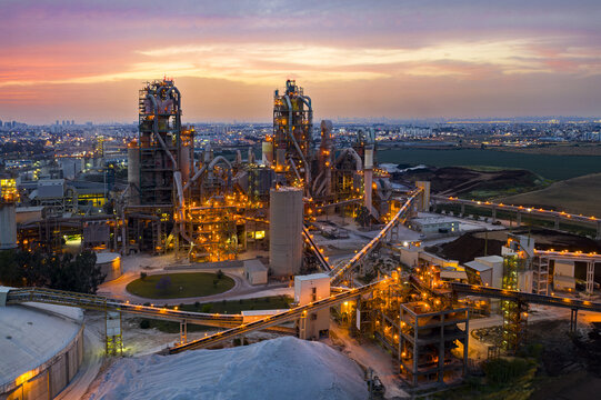 Aerial view of the Nesher cement factory illuminated at sunset with industrial silos, conveyor belts, and the city skyline in the background in Ramla, Center District, Israel.