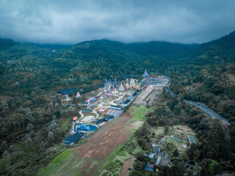 Aerial view of Hibisc Fantasy Puncak amusement park with a ferris wheel and colorful buildings nestled in lush green mountains under a cloudy sky Cisarua, West Java, Indonesia.