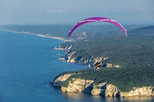 Aerial view of a paramotor pilot gliding above the Cilingoz cliffs, dense green forests, and the deep blue sea along the coast of Catalca, Istanbul, Turkey.