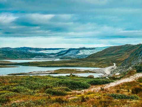 View of the vast landscape featuring a distant glacier, calm lake, rolling tundra hills, and a winding dirt road under a dramatic cloudy sky, Greenland.