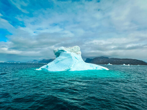 View of a large blue iceberg floating in the cold ocean with distant mountains under a cloudy sky in Greenland.