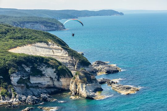 Aerial view of a paraglider soaring over the rugged coastline of Yalikoy, featuring dramatic cliffs, turquoise sea, and lush green hills in Catalca, Istanbul, Turkey.