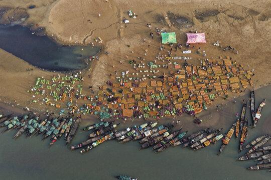 Aerial view of a bustling riverbank market with numerous wooden boats moored along the shore and vendors selling fresh produce on the sandy bank in Sylhet, Sylhet Division, Bangladesh.