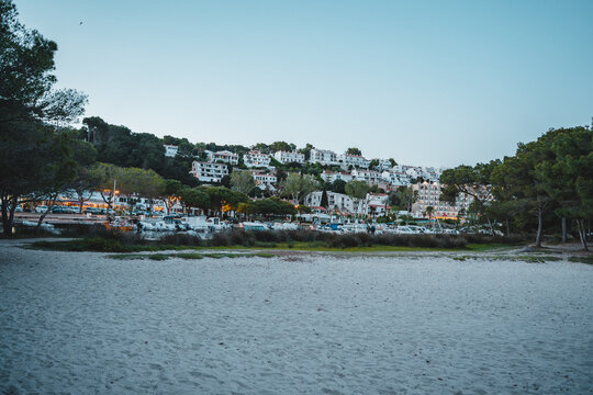 View of the sandy beach and white resort buildings nestled on the hillside with pine trees and boats in the harbor at Cala Galdana, Illes Balears, Spain.