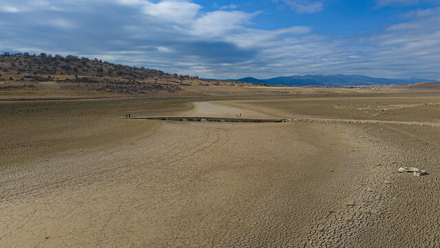 Aerial view of the dried-up bed of Apa Dam with cracked earth, a small stone bridge, and distant hills under a blue sky in Cumra, Konya, Turkiye.
