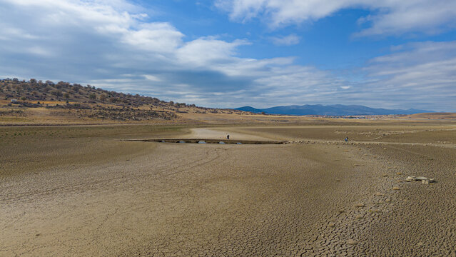 Aerial view of the dried-up bed of Apa Dam with cracked earth, a small stone bridge, and distant hills under a blue sky in Cumra, Konya, Turkiye.