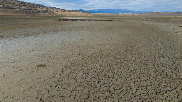 Aerial view of the dried-up bed of Apa Dam with extensive cracked earth patterns, a small stone bridge in the distance, and arid hills in Cumra, Konya, Turkiye.